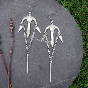 May include: A pair of silver earrings with a dangling arrow design. Each earring features a small, pointed arrow pendant hanging from a chain.