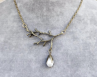 May include: A brass-coloured necklace featuring a branch design with delicate twigs. A pear-shaped pendant with a white pearl hangs from the branch. The necklace is displayed on a grey mannequin.