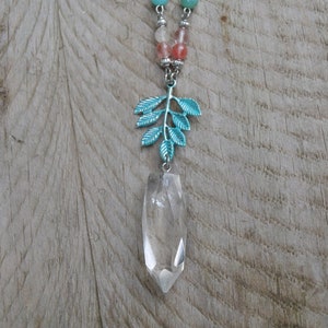 May include: A silver necklace with a clear crystal pendant and a teal leaf charm. The necklace has a chain with colourful beads in shades of blue, pink, and brown. The pendant is a clear, elongated crystal.