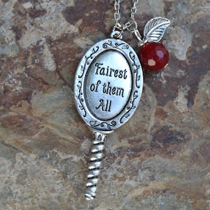 Peut inclure: Un collier argenté avec un pendentif ovale portant l'inscription "Fairest of them All". Une perle rouge et une breloque en forme de feuille pendent de la chaîne. Le pendentif a une poignée torsadée. Le collier est présenté sur une surface de pierre grise texturée.