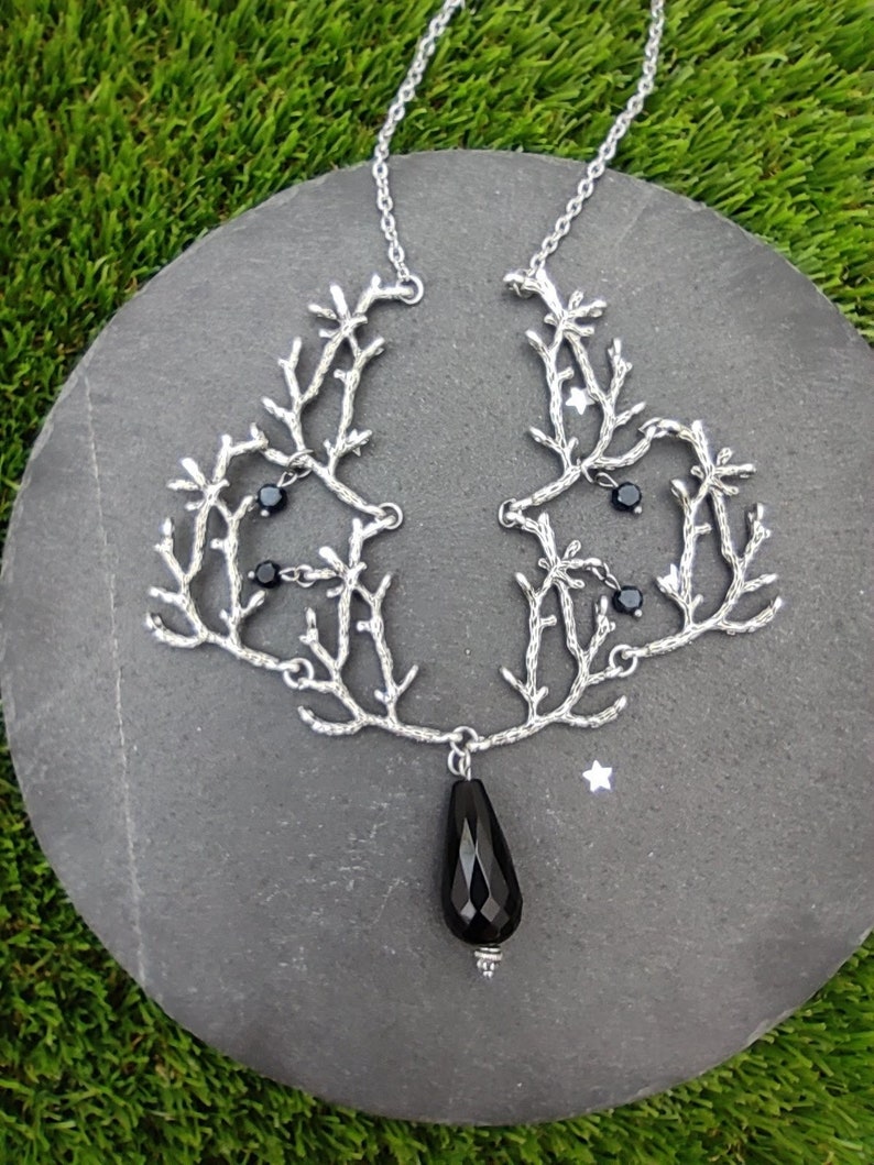 May include: A silver-toned necklace with a branch-like design, adorned with small black beads and a large, teardrop-shaped black gemstone pendant. The necklace is displayed on a gray stone against a backdrop of green grass.