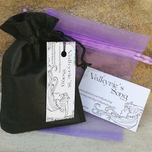 May include: A black drawstring bag with a tag and a business card, both displaying the text "Valkyrie's Song" in an elegant font. A sheer purple bag is visible behind the black bag. The tag and card feature a line drawing of a woman's face.