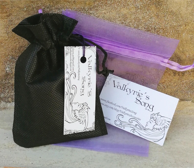 May include: A black drawstring bag with a tag and a business card, both displaying the text "Valkyrie's Song" in an elegant font. A sheer purple bag is visible behind the black bag. The tag and card feature a line drawing of a woman's face.