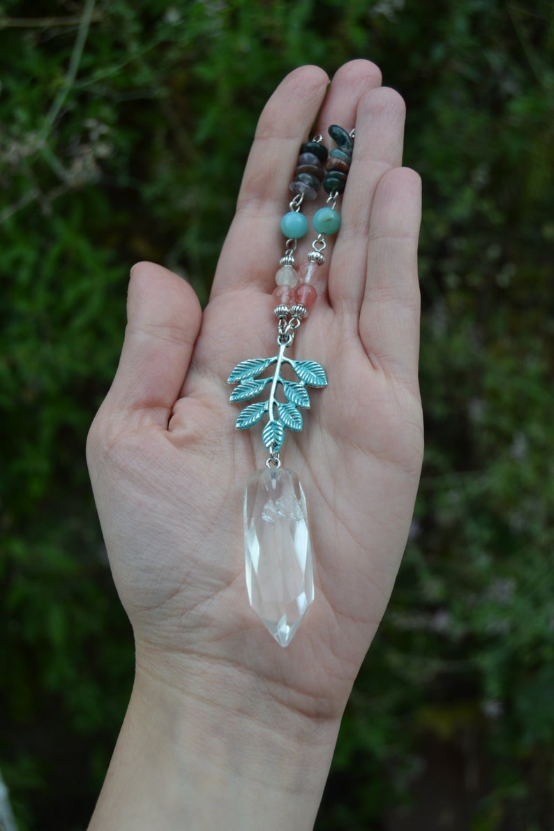May include: A clear quartz crystal pendant necklace with a silver-coloured leaf charm and colourful beaded accents. The pendant is suspended from a chain with turquoise, pink, and brown beads. The leaf charm has a teal finish.