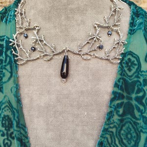 May include: A silver-toned necklace with a branch-like design, featuring small dark beads and a black teardrop pendant. The necklace is displayed on a neutral background, with the brand name "VALKYRIESSONG" visible.