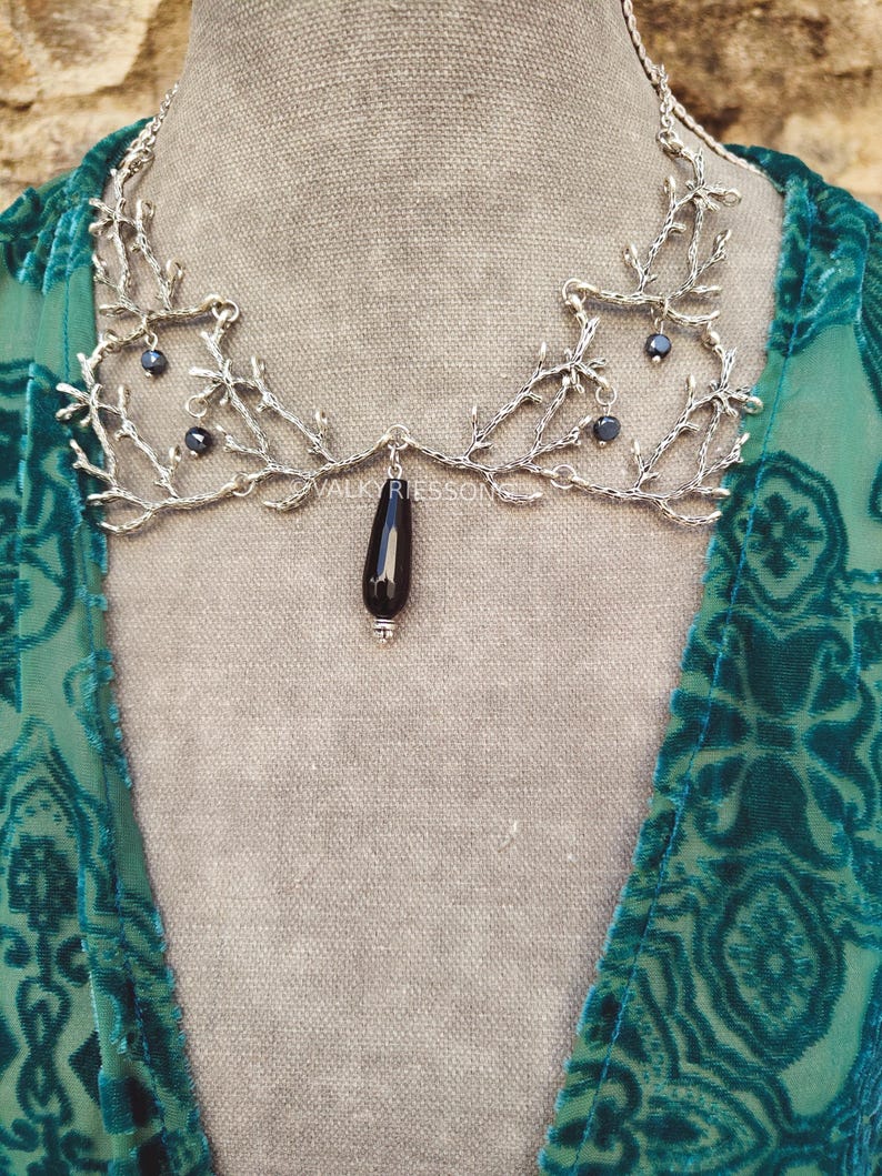 May include: A silver-toned necklace with a branch-like design, featuring small dark beads and a black teardrop pendant. The necklace is displayed on a neutral background, with the brand name "VALKYRIESSONG" visible.
