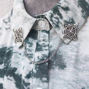 May include: Silver collar pin with a Celtic knot design. The pin is attached to a silver chain and is worn on a white and green tie-dye shirt.