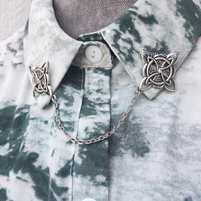 May include: Silver collar pin with a Celtic knot design. The pin is attached to a silver chain and is worn on a white and green tie-dye shirt.