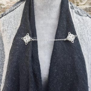 May include: A silver chain scarf holder with two decorative Celtic knot designs. The chain is attached to the knots, which are used to hold a scarf in place.
