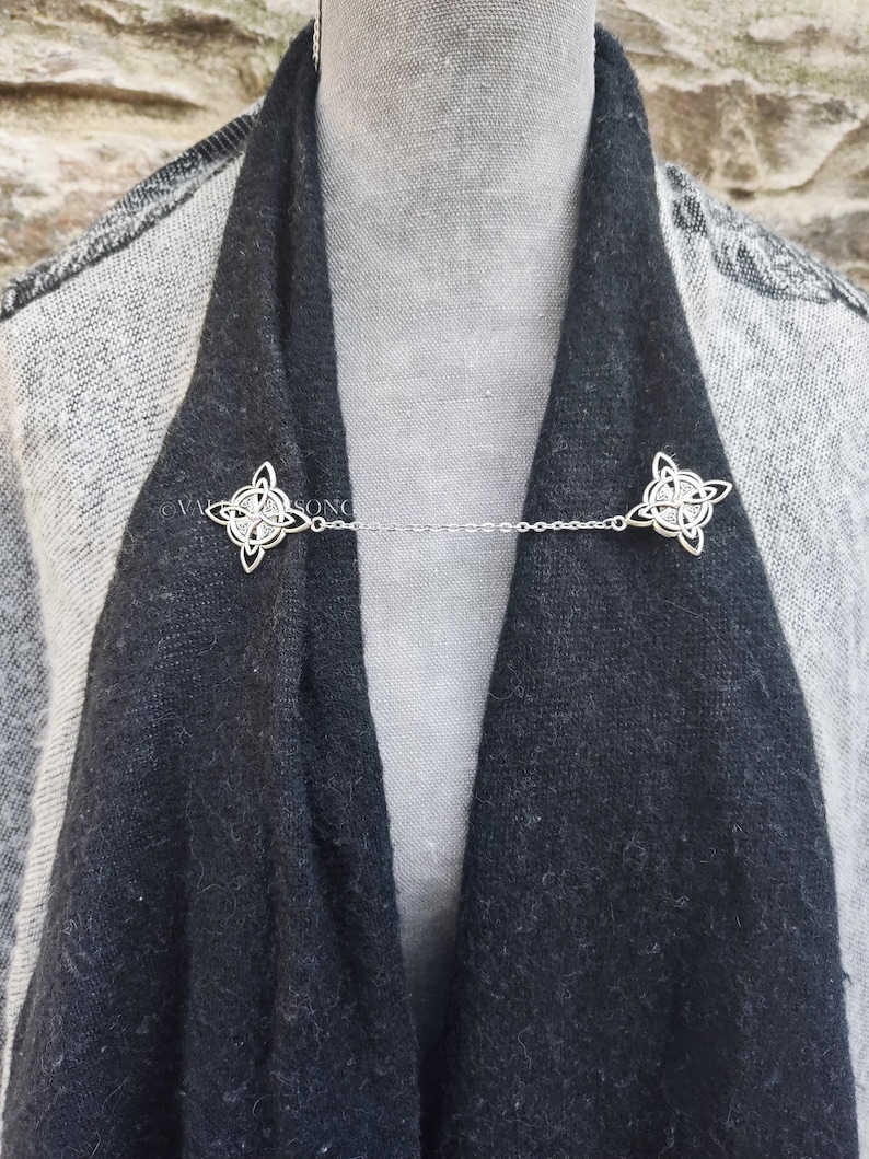 May include: A silver chain scarf holder with two decorative Celtic knot designs. The chain is attached to the knots, which are used to hold a scarf in place.