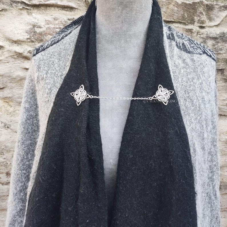 May include: A silver chain with two Celtic knot designs on each end. The chain is being used to hold a black scarf together.