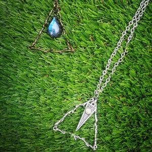 May include: Two silver triangle necklaces with gemstone pendants. The necklace on the left has a blue gemstone and the necklace on the right has a clear gemstone.