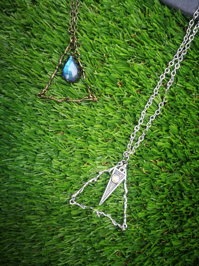 May include: Two silver triangle necklaces with gemstone pendants. The necklace on the left has a blue gemstone and the necklace on the right has a clear gemstone.