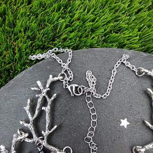 May include: A silver-toned necklace with a branch-like design. The necklace features small black beads and a chain with an adjustable length. The jewelry is displayed on a dark gray stone surface against a backdrop of green artificial grass.