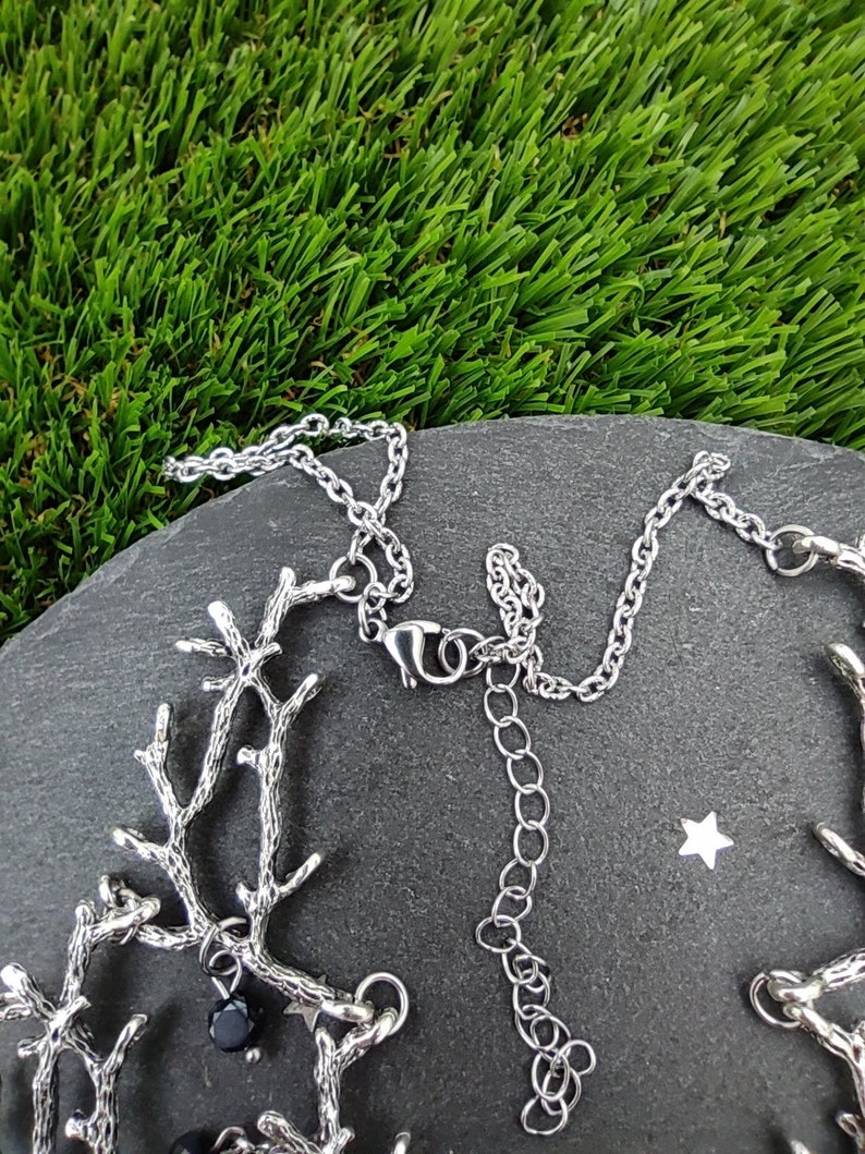 May include: A silver-toned necklace with a branch-like design. The necklace features small black beads and a chain with an adjustable length. The jewelry is displayed on a dark gray stone surface against a backdrop of green artificial grass.