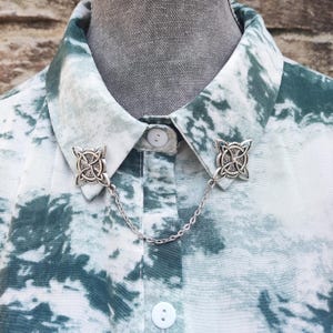 May include: A silver collar pin with a Celtic knot design. The pin is attached to a silver chain and is worn on a white shirt with a green and white pattern.