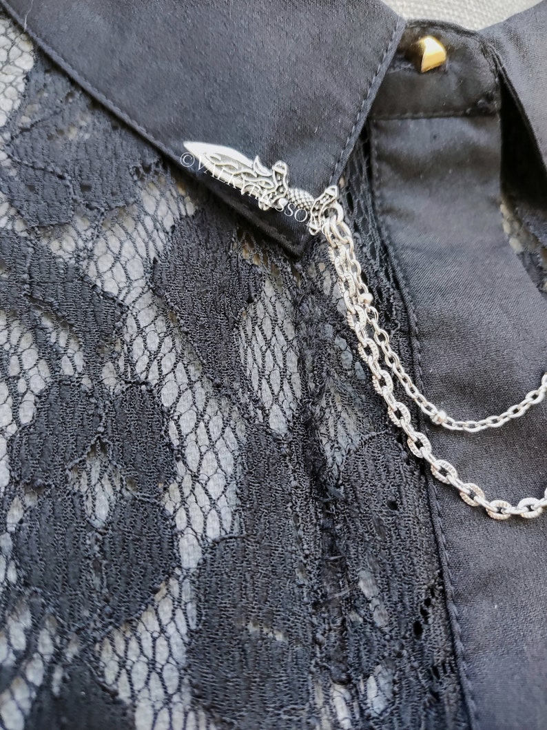 K&ouml;nnte beinhalten: Ein schwarzes Hemd mit einem Spitzeneinsatz und einem festen Kragen, verziert mit einer silberfarbenen Metallbrosche in Dolchform. Die Brosche ist mit zwei feinen silbernen Ketten verbunden, die &uuml;ber die Vorderseite des Kleidungsst&uuml;cks h&auml;ngen.