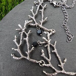 May include: Silver-toned necklace featuring a branch-like design with small black beads. The necklace is displayed on a dark gray stone surface, with green grass visible in the background. The chain is adjustable.