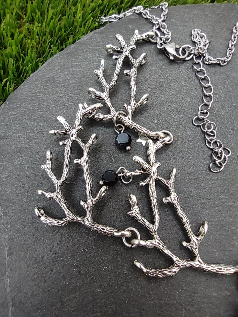 May include: Silver-toned necklace featuring a branch-like design with small black beads. The necklace is displayed on a dark gray stone surface, with green grass visible in the background. The chain is adjustable.