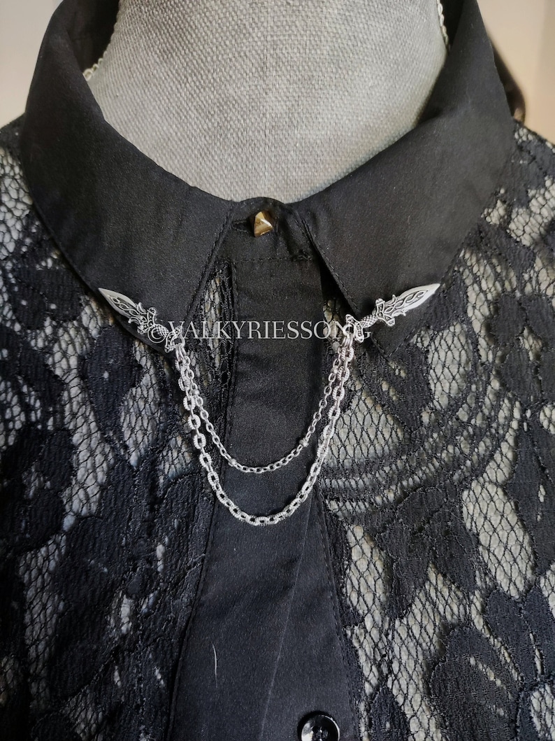 May include: A black collared shirt with a lace overlay. The collar is fastened with a silver chain and two silver sword-shaped pins.