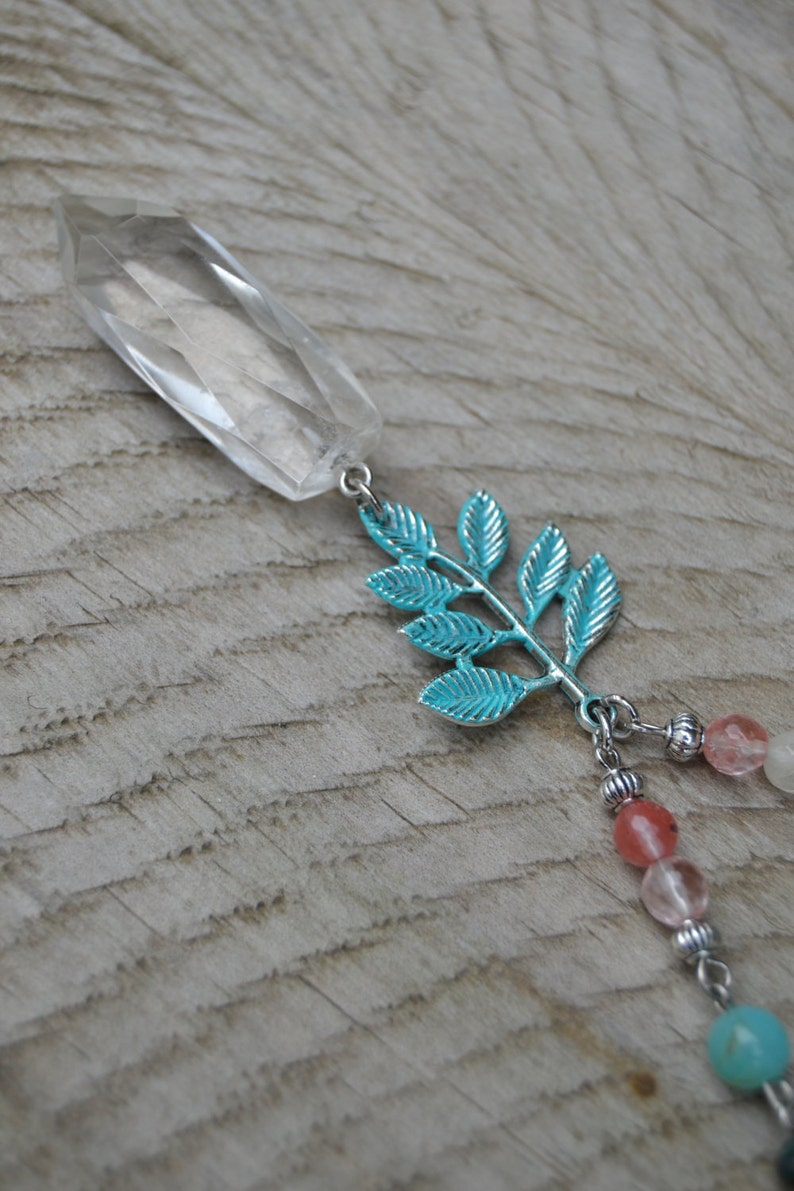 May include: A clear, faceted crystal pendant with a silver-colored metal leaf design and a strand of colorful beads. The leaf is painted a bright turquoise color. The beads are pink, white, and turquoise.