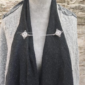 May include: A silver chain scarf clip with two Celtic knot designs. The clip is holding a black and gray scarf together.