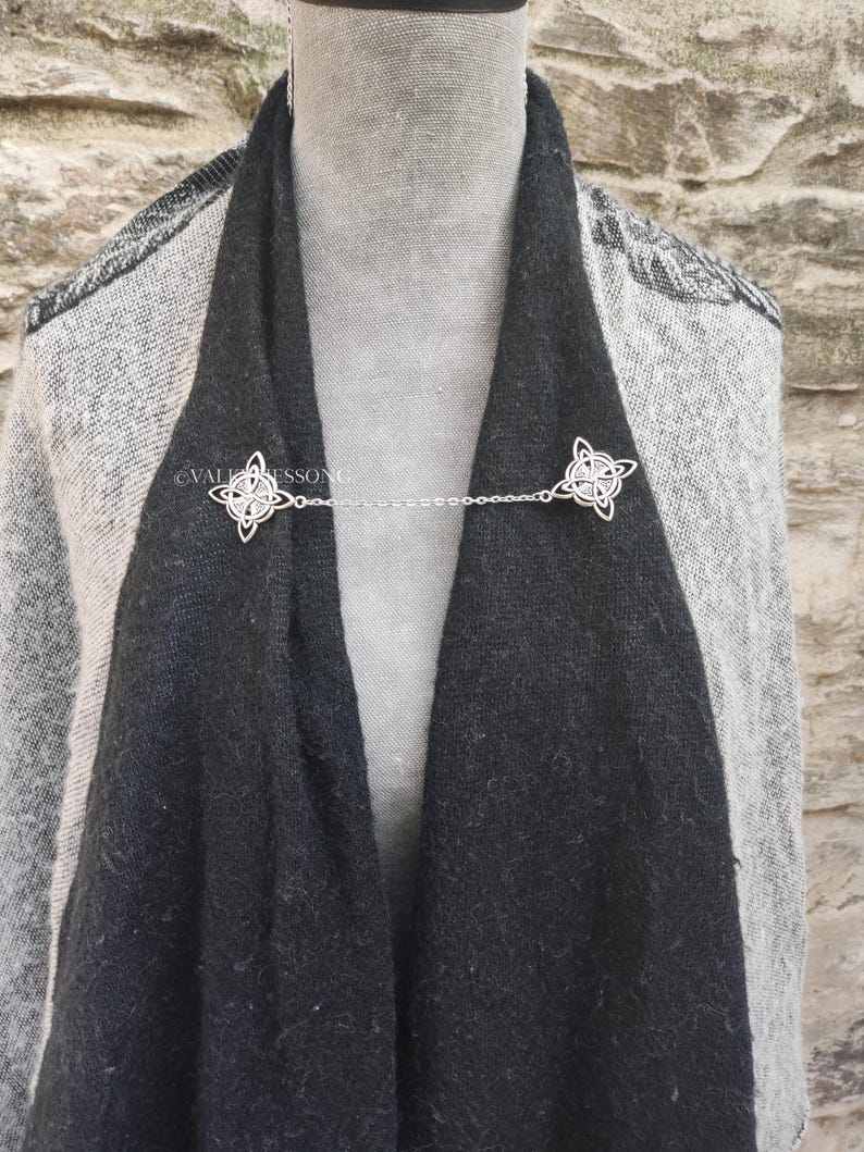 May include: A silver chain scarf clip with two Celtic knot designs. The clip is holding a black and gray scarf together.