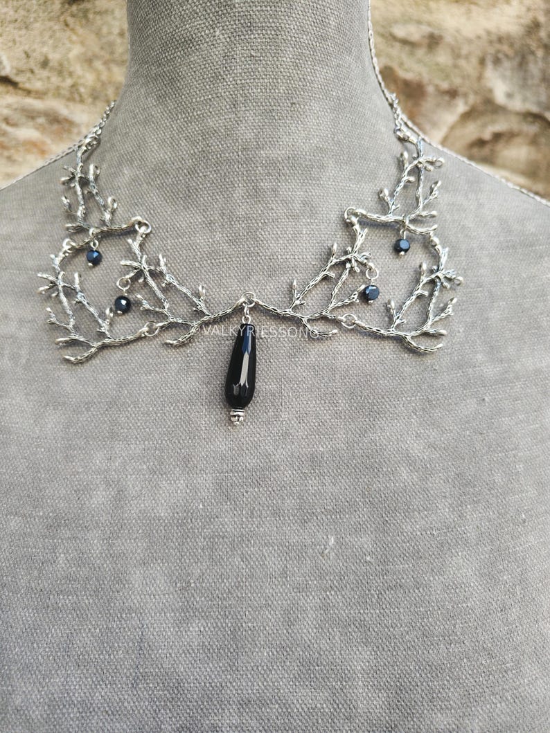 May include: Silver-toned statement necklace with a branch-like design. The necklace features small, dark blue beads and a black teardrop-shaped pendant. The necklace is displayed on a mannequin.
