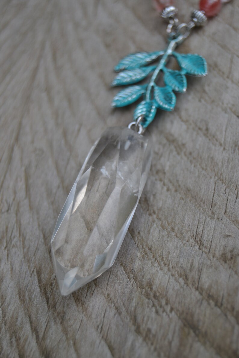 May include: A clear quartz crystal pendant necklace with a faceted, elongated shape. The pendant hangs from a silver chain with a turquoise-coloured leaf design. The necklace features a strand of pink and white beads.