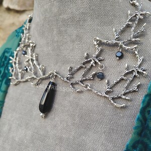 May include: A silver-toned necklace with a branch-like design. The necklace features small, dark beads and a large, black teardrop-shaped pendant. The necklace is displayed on a gray surface.