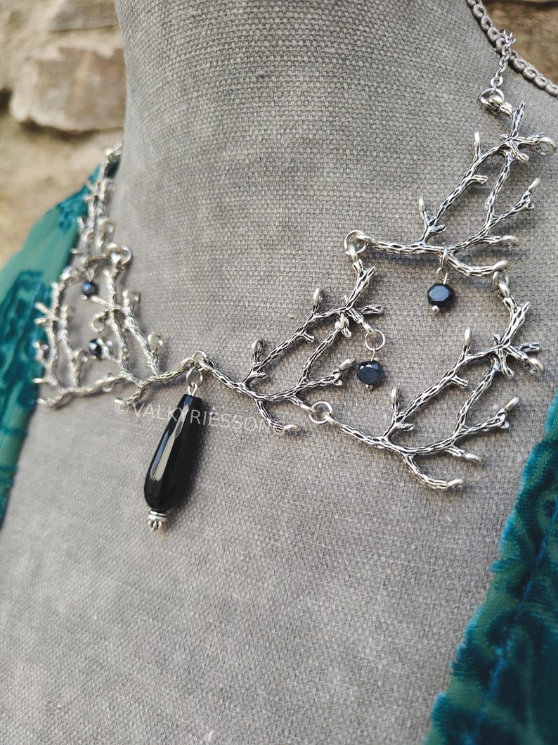 May include: A silver-toned necklace with a branch-like design. The necklace features small, dark beads and a large, black teardrop-shaped pendant. The necklace is displayed on a gray surface.