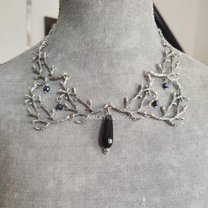 May include: Silver-toned necklace with a branch-like design, adorned with small dark blue beads. A black teardrop-shaped pendant hangs from the center. The necklace is displayed on a gray mannequin.