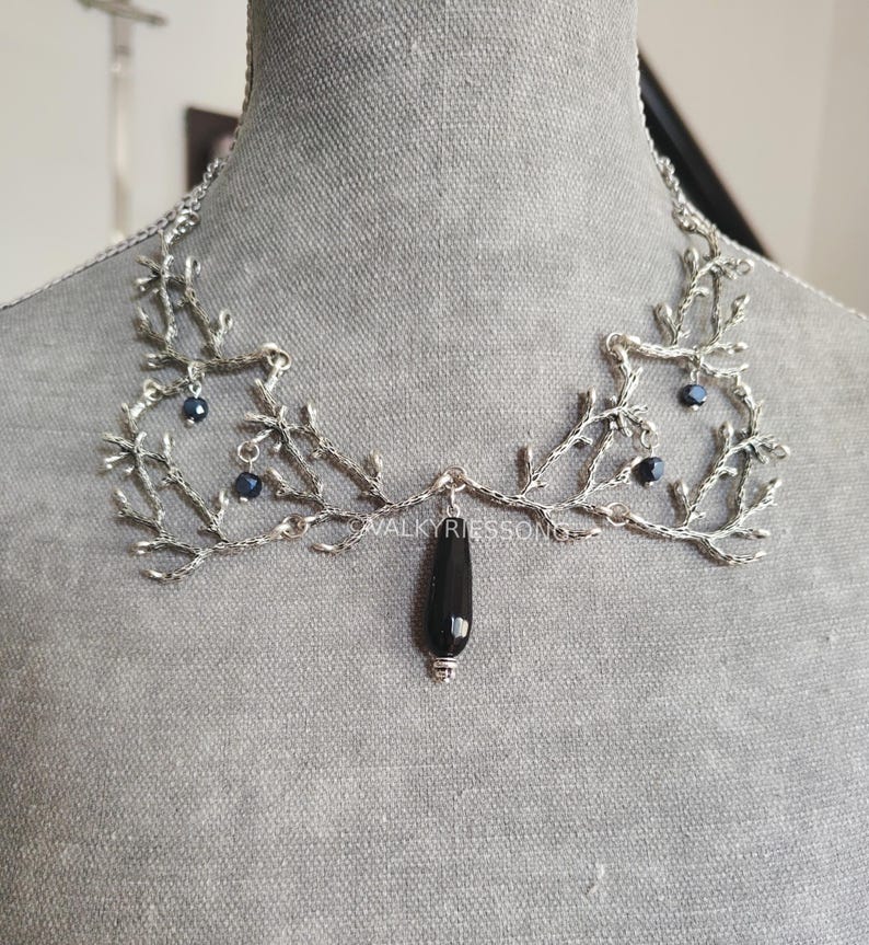 May include: Silver-toned necklace with a branch-like design, adorned with small dark blue beads. A black teardrop-shaped pendant hangs from the center. The necklace is displayed on a gray mannequin.