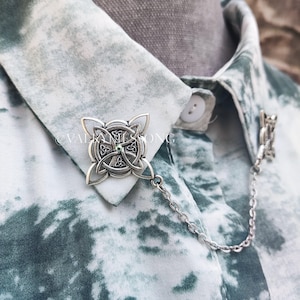 May include: A silver collar pin with a Celtic knot design and a small green gemstone in the center. The pin is attached to a silver chain.