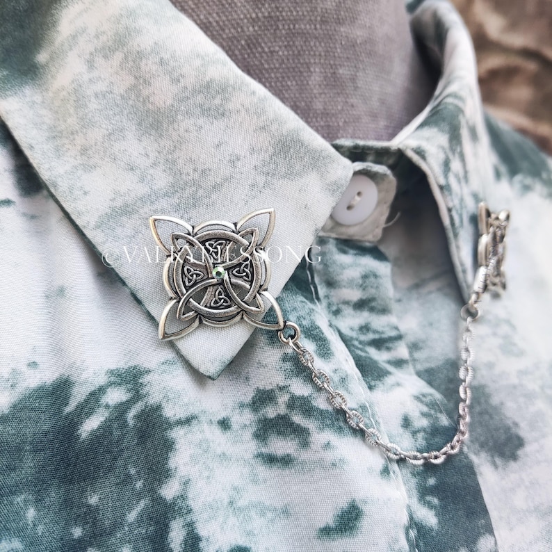 May include: A silver collar pin with a Celtic knot design and a small green gemstone in the center. The pin is attached to a silver chain.