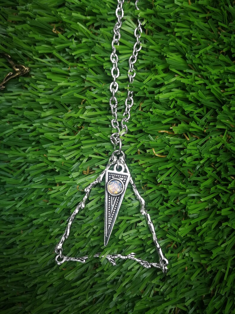 May include: A silver necklace with a triangular pendant featuring a small iridescent gemstone. The pendant is surrounded by a delicate, intricate silver design.