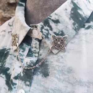 May include: A silver collar pin with a Celtic knot design. The pin is attached to a silver chain and is worn on a white and green patterned shirt.
