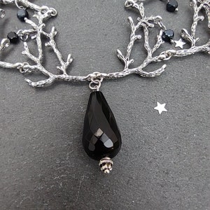 May include: A silver-toned necklace with a branch-like design. The necklace features a large, faceted black teardrop pendant and small black beads. The background is a dark gray surface with small star-shaped accents.
