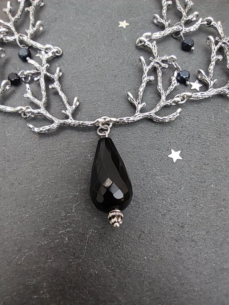May include: A silver-toned necklace with a branch-like design. The necklace features a large, faceted black teardrop pendant and small black beads. The background is a dark gray surface with small star-shaped accents.