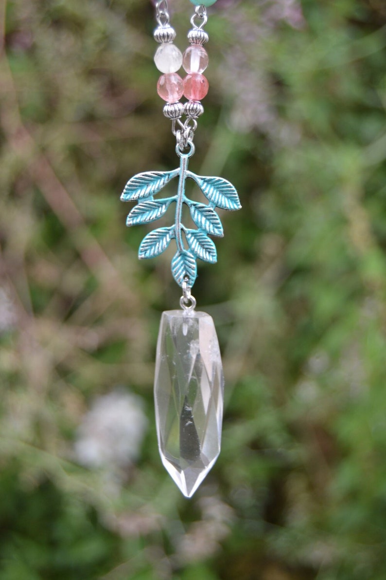 May include: A clear quartz crystal pendant with a faceted design hangs from a beaded chain. The pendant is suspended from a teal-coloured metal leaf charm. The chain features pink and white beads.