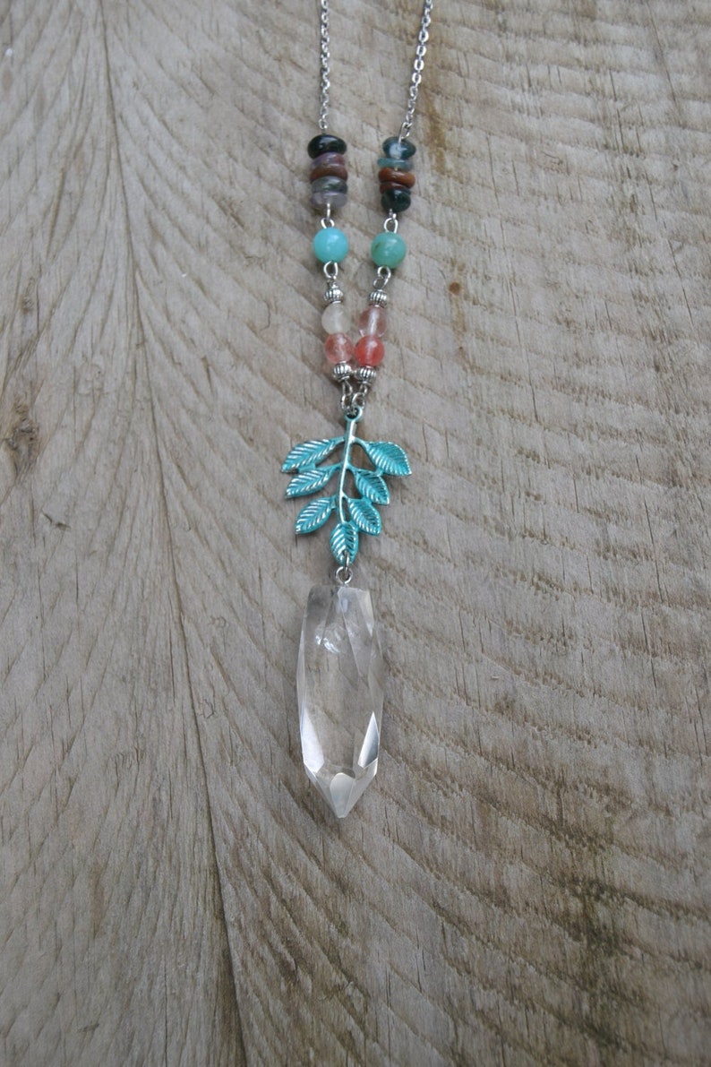 May include: A silver necklace with a clear crystal pendant and a teal leaf charm. The necklace has a chain with colourful beads in shades of blue, pink, and brown. The pendant is a clear, elongated crystal.