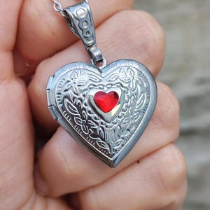 May include: A silver-toned heart-shaped locket pendant on a matching chain. The locket features an intricate embossed floral pattern and a small, faceted red heart-shaped stone set in the center. The text VALKYRIESSONG is engraved at the top.