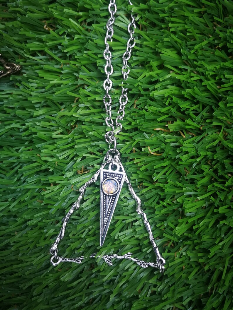 May include: A silver necklace with a triangular pendant featuring a small, iridescent gemstone. The pendant is attached to a delicate chain and hangs from a simple, open triangle frame.