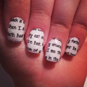 May include: Close-up of a hand with white nail polish and black text from a book on each nail. The text reads "Lack a", "Then I d", "Hath had", "ry cat a", "on her, b", "y unwor", "I see th", "tom of a", "in the", "I fath", "yer be", "not ma".