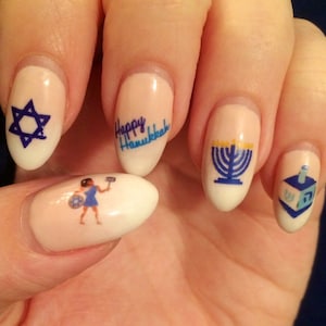 May include: Close-up of a hand with five fingernails painted white and decorated with blue and gold Hanukkah-themed nail art. The designs include a Star of David, a menorah, a dreidel, and the words "Happy Hanukkah".