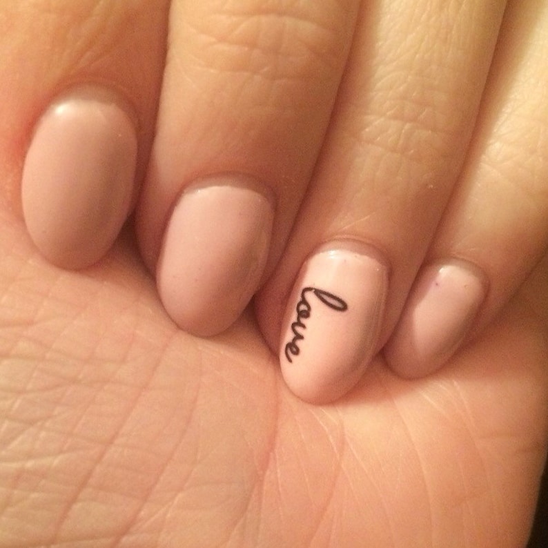 May include: Close-up of a hand with four nude-colored nails. The ring finger nail has the word "love" written on it in black.