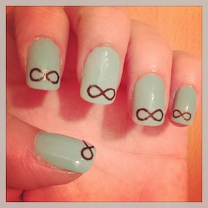 Peut inclure: Gros plan d'une main avec un vernis à ongles bleu clair et des symboles d'infini peints sur chaque ongle.