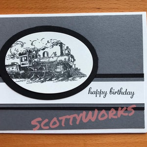 May include: A handmade birthday card featuring a black and white illustration of a vintage steam train inside a black oval. The card has a gray and white layered design with the words "happy birthday" in black script.