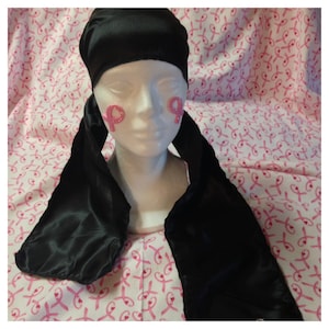 May include: Black satin hair scarf with long tails, tied around a white mannequin head. The scarf is on a pink background with pink ribbon designs.