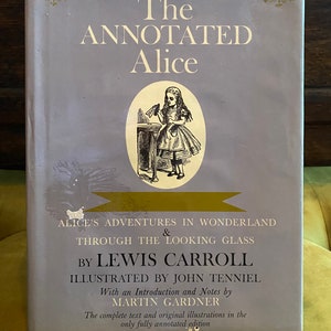 May include: A vintage book cover with a purple background featuring the title "The Annotated Alice" in gold lettering. The cover also includes an illustration of Alice from "Alice's Adventures in Wonderland" by Lewis Carroll, illustrated by John Tenniel. The book is the only fully annotated edition with an introduction and notes by Martin Gardner.
